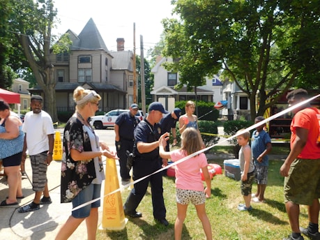 A group of people, including adults and children, are gathered outdoors in a neighborhood setting. Some individuals are interacting with two uniformed police officers. The scene is colorful and lively, suggesting a community event. Residential houses with trees are visible in the background.