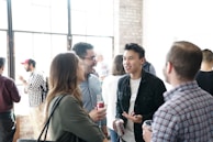 A group of people is gathered in a well-lit room with large windows and exposed brick walls. They appear to be engaged in conversation, holding drinks. The atmosphere is casual and sociable.
