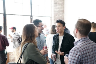 A group of people is gathered in a well-lit room with large windows and exposed brick walls. They appear to be engaged in conversation, holding drinks. The atmosphere is casual and sociable.