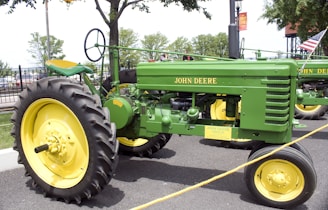 A classic John Deere tractor is parked on a paved surface, surrounded by a rope barrier. The tractor is painted in the traditional green and yellow colors. It has large rear wheels and smaller front wheels, with a visible seat and steering wheel. Trees and a section of fence are visible in the background.
