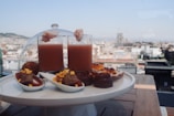 A selection of gourmet snacks is elegantly arranged on a white platter with a cityscape view in the background. Two tall glasses contain a reddish beverage topped with shrimp. Various bite-sized appetizers, including chocolates and other sweets, are displayed around the glasses.