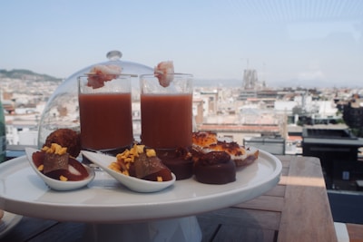 A selection of gourmet snacks is elegantly arranged on a white platter with a cityscape view in the background. Two tall glasses contain a reddish beverage topped with shrimp. Various bite-sized appetizers, including chocolates and other sweets, are displayed around the glasses.