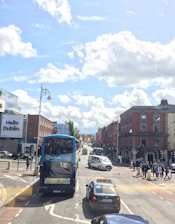 A busy urban street scene in Dublin with a blue double-decker bus and a taxi among other vehicles at a traffic light. Pedestrians are crossing the street, and red brick buildings line the road. Bright billboard advertising and a clear blue sky with scattered clouds are visible.