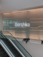 An illuminated sign displaying 'Bershka' is mounted on a modern glass wall. Below the sign is an escalator with potted plants placed along the rail. The setting appears to be inside a retail shopping environment with ambient lighting creating a sleek and contemporary atmosphere.