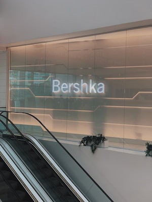 An illuminated sign displaying 'Bershka' is mounted on a modern glass wall. Below the sign is an escalator with potted plants placed along the rail. The setting appears to be inside a retail shopping environment with ambient lighting creating a sleek and contemporary atmosphere.