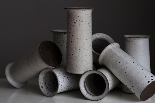 A collection of handmade ceramic vases with a speckled glaze are artfully arranged against a dark background. They are cylindrical with a minimalistic design and varying shades of gray.
