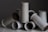 A collection of handmade ceramic vases with a speckled glaze are artfully arranged against a dark background. They are cylindrical with a minimalistic design and varying shades of gray.