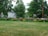 A lush green lawn is bordered by vibrant flowerbeds full of colorful blooms, including pink, red, and orange flowers. Large leafy trees and a few houses frame the background, creating a serene residential scene.
