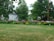 A lush green lawn is bordered by vibrant flowerbeds full of colorful blooms, including pink, red, and orange flowers. Large leafy trees and a few houses frame the background, creating a serene residential scene.