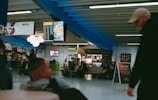 The image depicts a busy indoor setting, likely an airport or shopping center, with people seated and walking around. An informational digital display and various advertisements are visible in the background. The area is well-lit, and there are signs indicating a shop and service center. The environment appears casual and bustling with activity, with a blue structural beam visible above.