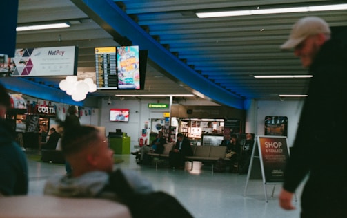 The image depicts a busy indoor setting, likely an airport or shopping center, with people seated and walking around. An informational digital display and various advertisements are visible in the background. The area is well-lit, and there are signs indicating a shop and service center. The environment appears casual and bustling with activity, with a blue structural beam visible above.