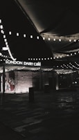A dimly lit outdoor area featuring the entrance of 'London Dairy Café', with decorative string lights hanging overhead. The brick wall has Arabic script painted on it, and there are shadows cast across the patterned ground.