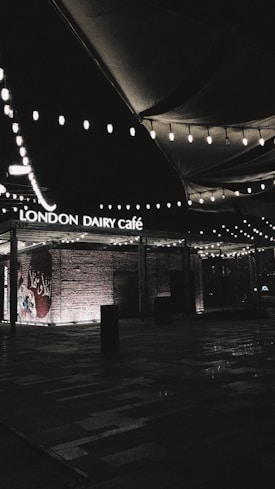 A dimly lit outdoor area featuring the entrance of 'London Dairy Café', with decorative string lights hanging overhead. The brick wall has Arabic script painted on it, and there are shadows cast across the patterned ground.