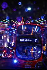 A red double-decker bus is driving through a city street at night, heading toward East Acton on route number 7. The street is festively decorated with colorful neon lights and spherical, glittering decorations. The bus's interior is illuminated, revealing several passengers on board. The background features lit-up buildings and other vehicles.