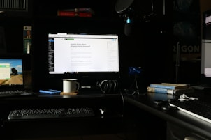 A workspace featuring multiple computer screens is set up on a dark desk. The main central monitor displays a webpage about blogging myths. To the left, a laptop shows a scenic desktop background, and another monitor on the right displays a webpage. A keyboard, headphones, a coffee mug, and a smartphone are placed on the desk. Books, including titles on Provence and psychology, are stacked next to a microphone setup.