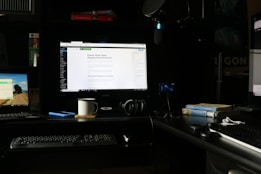 A workspace featuring multiple computer screens is set up on a dark desk. The main central monitor displays a webpage about blogging myths. To the left, a laptop shows a scenic desktop background, and another monitor on the right displays a webpage. A keyboard, headphones, a coffee mug, and a smartphone are placed on the desk. Books, including titles on Provence and psychology, are stacked next to a microphone setup.