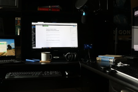 A workspace featuring multiple computer screens is set up on a dark desk. The main central monitor displays a webpage about blogging myths. To the left, a laptop shows a scenic desktop background, and another monitor on the right displays a webpage. A keyboard, headphones, a coffee mug, and a smartphone are placed on the desk. Books, including titles on Provence and psychology, are stacked next to a microphone setup.