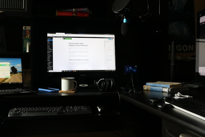 A workspace featuring multiple computer screens is set up on a dark desk. The main central monitor displays a webpage about blogging myths. To the left, a laptop shows a scenic desktop background, and another monitor on the right displays a webpage. A keyboard, headphones, a coffee mug, and a smartphone are placed on the desk. Books, including titles on Provence and psychology, are stacked next to a microphone setup.