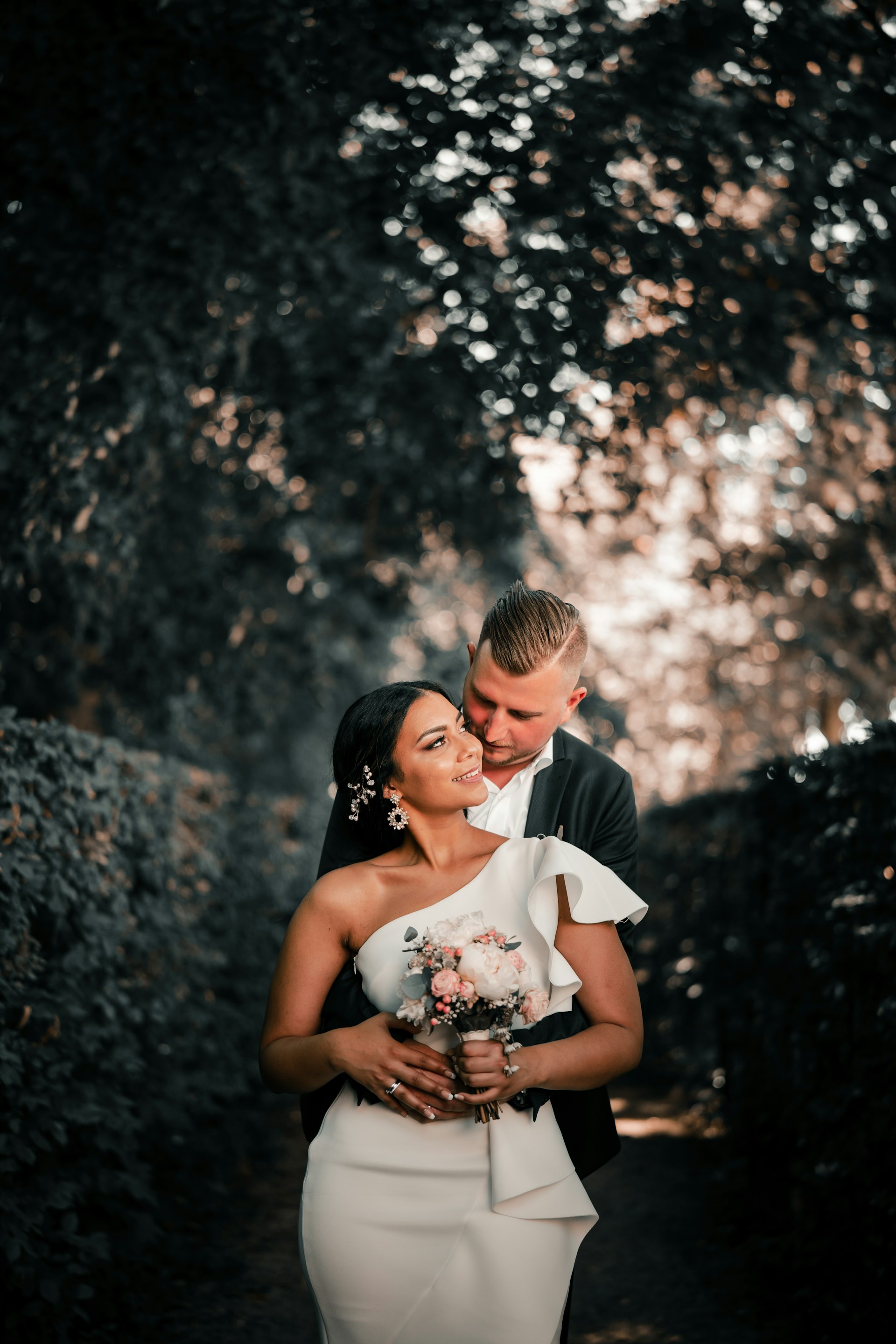 man hugging woman in white dress from behind between plants