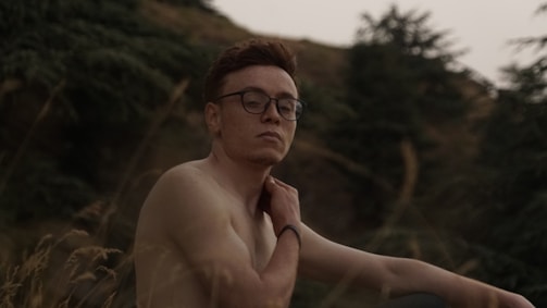 A person with short hair and glasses is sitting outdoors in a natural setting. The background features trees and a hillside, adding to a serene atmosphere. The person appears relaxed and contemplative.