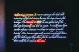 A neon art installation displays text about Robert Henry Lawrence Jr., an astronaut who died in a flight accident in 1967. The text highlights his tragic death and the racist responses that followed. Various neon colors are used to emphasize parts of the text, creating a vivid visual effect against a dark background.