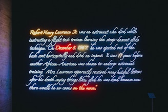 A neon art installation displays text about Robert Henry Lawrence Jr., an astronaut who died in a flight accident in 1967. The text highlights his tragic death and the racist responses that followed. Various neon colors are used to emphasize parts of the text, creating a vivid visual effect against a dark background.