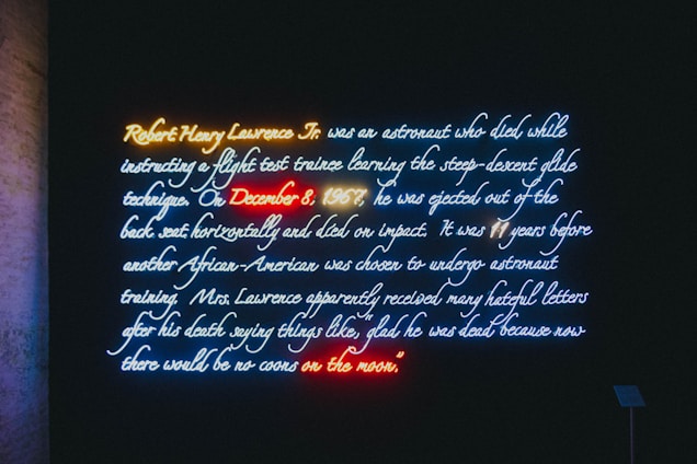 A neon art installation displays text about Robert Henry Lawrence Jr., an astronaut who died in a flight accident in 1967. The text highlights his tragic death and the racist responses that followed. Various neon colors are used to emphasize parts of the text, creating a vivid visual effect against a dark background.