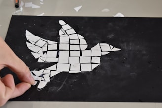 A hand is assembling white pieces of paper or tiles in the shape of a bird on a black background. The mosaic-like creation is composed of various geometric shapes.