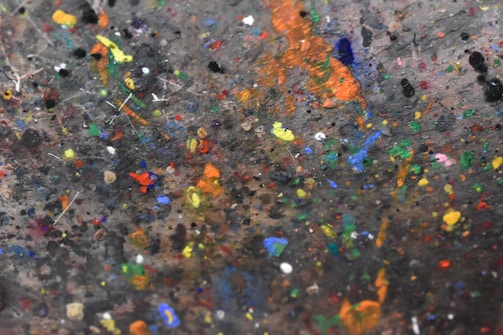 A chaotic explosion of various colored paint splatters covers a surface. The spots and smears range from bright oranges and yellows to deep blues and greens, creating a vibrant and eclectic mix. The surface appears worn and textured, suggesting layers of paint accumulated over time.