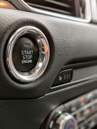 A close-up view of a car's start-stop engine button located on a leather-textured dashboard. The button has a metallic ring around it, and nearby, there's a panel with seating controls.