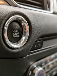 A close-up view of a car's start-stop engine button located on a leather-textured dashboard. The button has a metallic ring around it, and nearby, there's a panel with seating controls.