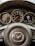 A close-up view of a car dashboard, featuring a steering wheel with a Mazda emblem in the center. Behind the steering wheel, several gauges display speed, fuel level, and other metrics.
