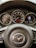 A close-up view of a car dashboard, featuring a steering wheel with a Mazda emblem in the center. Behind the steering wheel, several gauges display speed, fuel level, and other metrics.