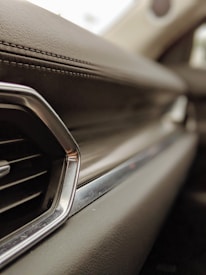 A close-up view of a car's interior focusing on the dashboard made of leather with detailed stitching. The image highlights a sleek air vent with metallic accents and a smooth, slightly reflective surface, all in soft focus, giving a sense of luxury.