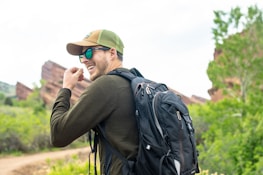 A person is outdoors, wearing a green cap, sunglasses, and a backpack, with a background of red rocks and lush greenery.