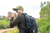 A person is outdoors, wearing a green cap, sunglasses, and a backpack, with a background of red rocks and lush greenery.