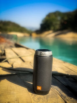 A black portable speaker is placed on a wooden surface with a blurred background that includes a serene body of water and greenery.