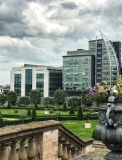 A modern urban garden with meticulously trimmed hedges and geometric designs, set against the backdrop of contemporary glass office buildings. The sky is overcast, giving a dramatic feel, while a decorative stone vase with vibrant purple and white flowers is visible in the foreground.
