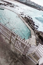 A coastal swimming pool with turquoise water is surrounded by a white picket fence and rocky formations. People are swimming in the pool, and beyond it, the ocean waves crash against the rocks. In the background, there is a sandy beach and houses lining the coast.