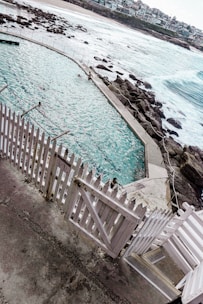 A coastal swimming pool with turquoise water is surrounded by a white picket fence and rocky formations. People are swimming in the pool, and beyond it, the ocean waves crash against the rocks. In the background, there is a sandy beach and houses lining the coast.