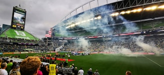 A packed football stadium with spectators observing a pre-game or halftime event on the field. Smoke drifts across the pitch, and an American flag is prominently displayed. The stands are filled with fans, and a large digital screen shows advertisements and game-related content.