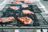 Several pieces of grilled meat resting on a barbecue grill, with a slightly charred appearance. Gray concrete blocks support the metal grill bars, and the background is softly blurred, highlighting the meat.