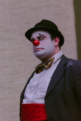 A person dressed as a clown with white face paint, a red nose, and exaggerated facial expressions is wearing a dark bowler hat, a bow tie, and a white shirt with a red sash. The background is a neutral beige color.