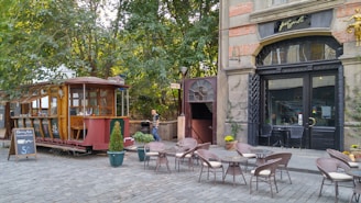 An outdoor cafe with wicker chairs and small round tables is arranged on a cobblestone street. A vintage tram car, repurposed as a part of the cafe, is parked nearby under green trees. A large building with an arched doorway and decorative elements serves as a backdrop. A chalkboard menu is displayed at the entrance.
