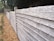 A wooden fence made up of horizontally stacked planks runs along a path lined with small green shrubs. The fence appears weathered with a rustic look. In the background, there are trees and a small structure that looks like a wooden shed or hut.