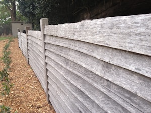A wooden fence made up of horizontally stacked planks runs along a path lined with small green shrubs. The fence appears weathered with a rustic look. In the background, there are trees and a small structure that looks like a wooden shed or hut.