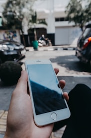 A person is holding a smartphone in one hand. The phone is turned off, showing a black screen. The background reveals an outdoor urban setting with parked cars, a motorcycle in the distance, trees, and a building.