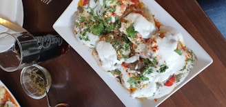 A rectangular white plate holds a dish covered in a creamy white sauce, garnished with fresh green herbs and sprinkled with red seasoning. The dish appears to include pieces of meat or vegetables covered in the sauce. Accompanying the dish on the table are a glass of cola with visible branding and a small bowl with a spoon containing a green sauce.