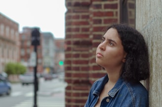 A person stands against a brick wall, gazing upwards with a contemplative expression. The background features an urban setting with blurred elements like buildings, a traffic light, and a street.