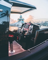 A luxurious yacht with a sleek, modern design is docked near a marina during sunset. The steering wheel and control panel of the yacht are visible, featuring polished metal and wood accents. The background shows another yacht and a building with a slightly vintage architecture. The scene is enhanced by the reflection of palm trees and the warm glow of the sunset.